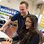 A smiling male educator standing over a female student who is laughing while working on a laptop in a classroom setting.