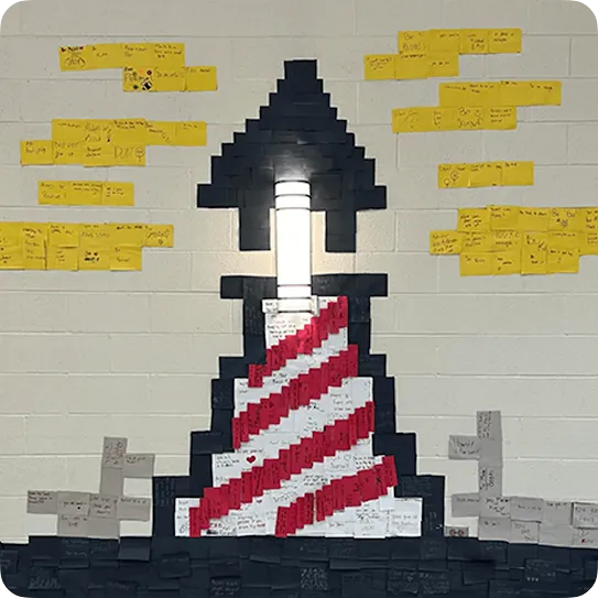 A creative wall mural of a red-and-white striped lighthouse built around an glowing light fixture using sticky notes, surrounded by yellow sticky notes arranged like clouds with handwritten inspirational messages.