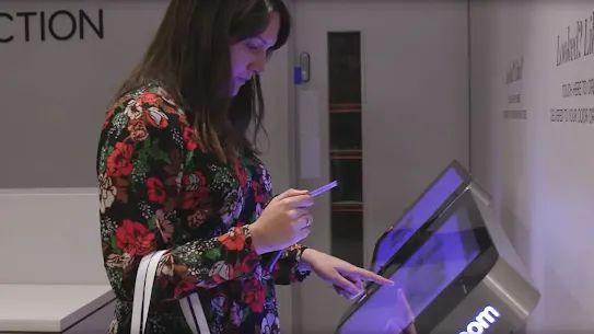 A side view of a woman using a touchscreen check-in kiosk while holding a blue pen or stylus.