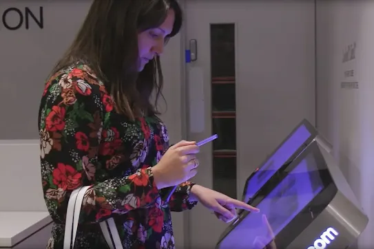 A side view of a woman using a touchscreen check-in kiosk while holding a blue pen or stylus.