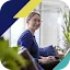 A young woman with glasses and a blue ribbed sweater smiles while working on a laptop in a bright, modern office with indoor plants. The image is framed by a stylized graphic overlay in navy blue, lime yellow, and teal. This photo illustrates the Google and Ipsos 'AI Works for America' poll, which explores how training and tools help employees become 'AI Fluent' in the modern economy.