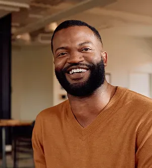 A smiling man with a beard wearing an orange V-neck sweater in a modern office or coworking space.