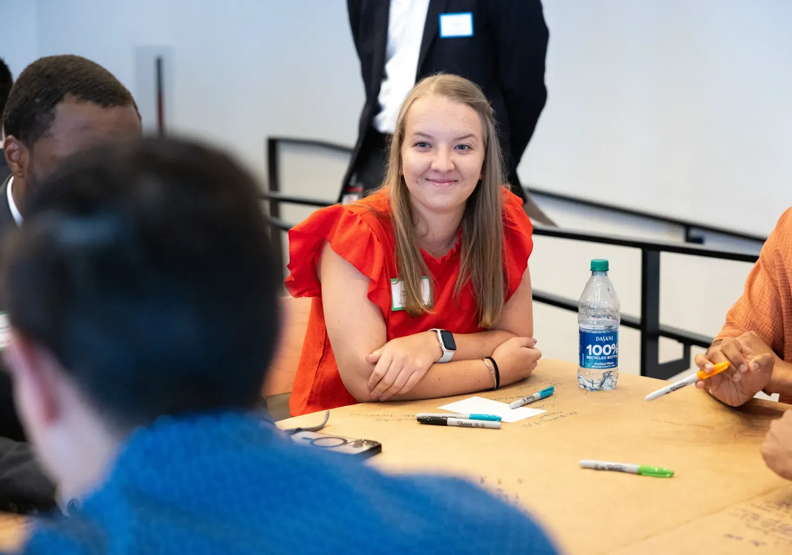 A young woman with blonde hair wearing a bright red ruffled top sits at a table during a workshop. She is smiling and looking toward someone off-camera. The table is covered in brown craft paper with handwritten notes and markers, and a bottle of Dasani water is visible.