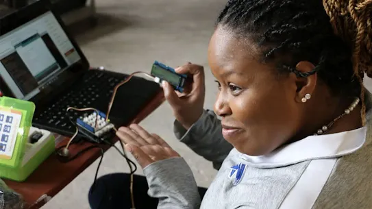 A student with braided hair smiling while working with a Thimble.io STEM kit. She is holding a small electronic component connected to a laptop and a circuit board on a desk in a classroom setting.