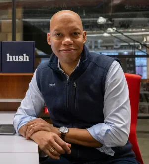 A professional portrait of a man in a navy blue vest sitting at a desk with "hush" branding visible in the background, representing a Michigan-based cybersecurity startup.