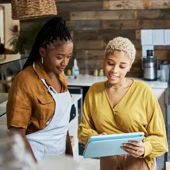 A cafe employee wearing an apron and a manager with short blonde hair looking at a blue tablet screen together behind a counter.