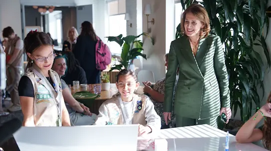 A woman in a green suit stands smiling while observing two young girls in scout vests working on a project at a table during an indoor event.