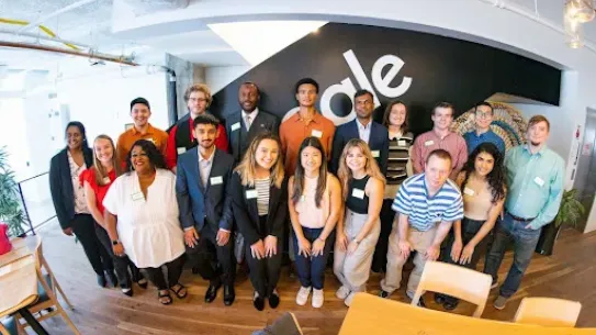 A wide-angle group photo of a diverse team of young professionals and students posing in a modern office. They are standing in front of a black wall featuring a large white Google "G" logo. The room has high ceilings, industrial pipes, and light wood flooring. Many individuals are wearing green name tags.