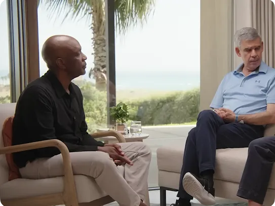 Two men engaged in a serious conversation while seated in comfortable armchairs. The man on the left is Black, wearing a black shirt and tan trousers; the man on the right is white with grey hair, wearing a light blue polo shirt. They are in a bright, modern room with a large window overlooking a coastal landscape and palm tree.