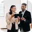 A smiling woman in a beige blazer and a man in a black suit standing together and looking at a digital tablet.
