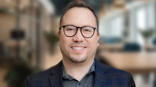 Professional headshot of AJ DeLeón, co-founder of Innovare, wearing glasses and a plaid blazer against a blurred office background.