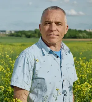 A portrait of a man with short graying hair wearing a light blue patterned button-down shirt, standing in a vast field of yellow wildflowers under a clear sky.