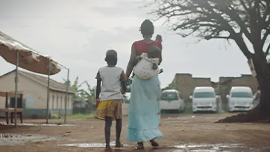 A woman carries her baby on her back and holds her daughter’s hands as they walk away along a dirty road.