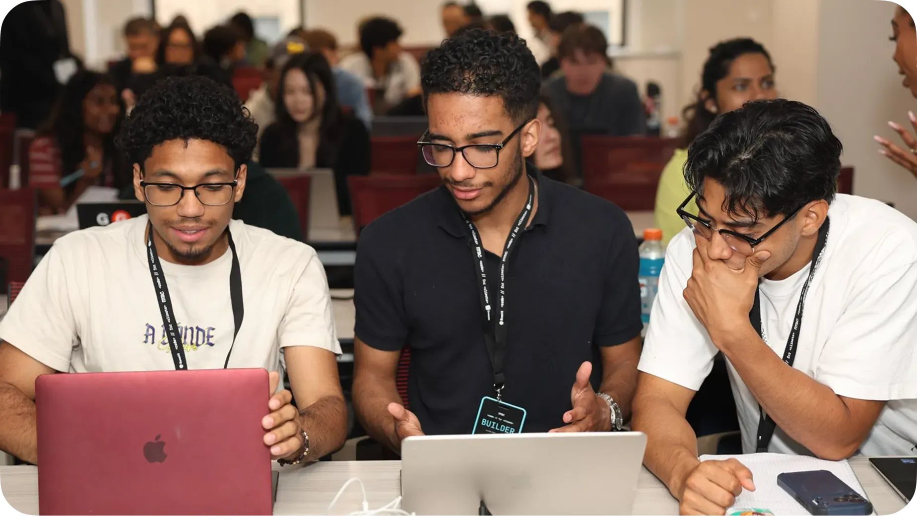Three young male students wearing glasses sit side-by-side at a desk collaborating over laptops during a workshop or tech event.