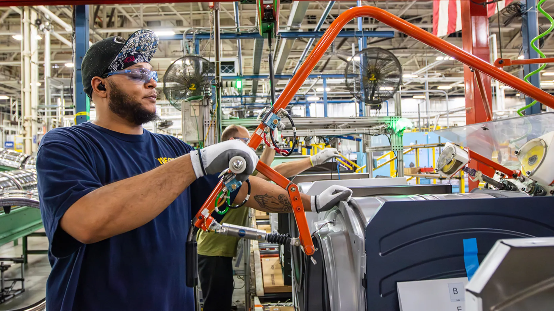 A male GE Appliances worker wearing safety glasses, a bandana under his hat, and protective gloves operates an orange ergonomic lift-assist tool on an assembly line in a manufacturing plant.