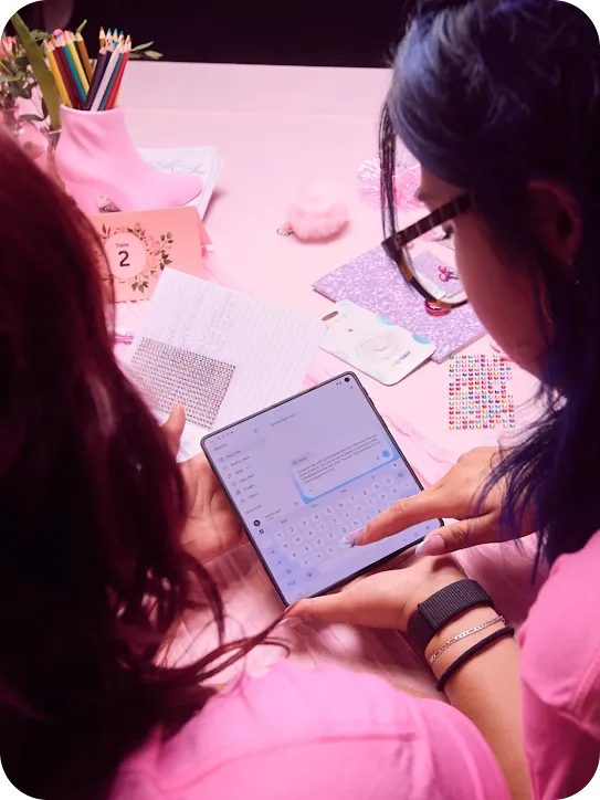 Over-the-shoulder view of two people using the Gemini app on a folding tablet at a table decorated with pink accessories, markers, and rhinestones.