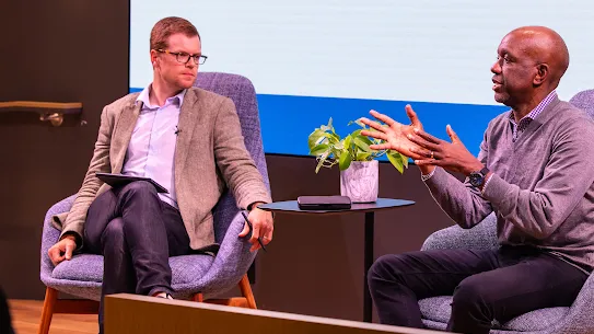 Ben Armstrong, Executive Director of the MIT Industrial Performance Center, and James Manyika, Google’s SVP of Research, Labs, Technology & Society, seated in blue armchairs during a panel discussion. James Manyika is speaking and gesturing with his hands while Ben Armstrong listens. A small potted plant sits on a table between them against a backdrop of a large presentation screen.