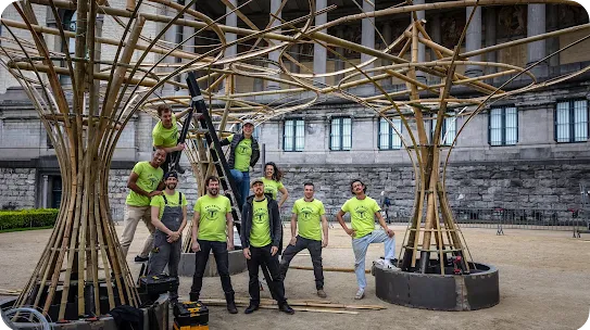 A group of eight construction or design team members wearing matching neon green t-shirts that say "THE TRIBE" pose together outside. They are standing around and climbing a ladder on a large, complex architectural pavilion made of curved bamboo poles. A historic stone building with columns stands in the background.