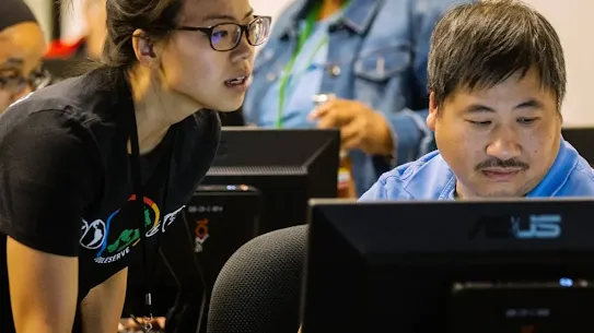 A candid photo from a digital skills training workshop. A woman wearing a Google badge and a black t-shirt leans in to assist a man seated at a computer monitor. Other participants are visible in the background of the classroom, illustrating the human impact of Google.org's future of work initiatives in Europe.