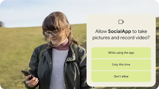 A person standing on a grassy hillslope looking at their Android phone. A graphic overlay seeking permission to allow an app to take pictures and record videos is placed on top. Permission options include allowing access while using the app, only this time or don’t allow.