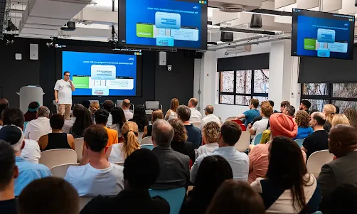 The host stands before a crowded event hall.
