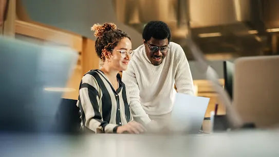 Two colleagues collaborating in a modern, brightly lit office space. A woman with curly hair tied up in a bun, wearing glasses and a striped button-down shirt, is seated at a desk typing on a laptop. A man wearing glasses and a cream-colored knit sweater stands beside her, leaning in slightly to look at the laptop screen with an engaged, smiling expression. The foreground and background show a softly blurred office environment.