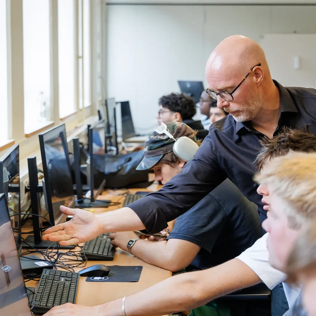 A teacher with glasses and a black shirt leans over to point at a computer monitor, instructing a male student