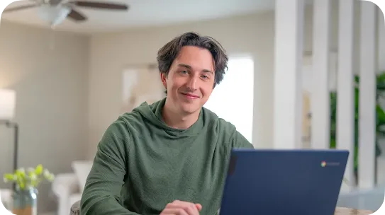 A young man with dark hair wearing a green textured hoodie sits at a table and smiles at the camera. A dark blue laptop is open in front of him in a brightly lit, modern home interior.