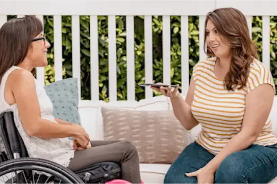 A woman sits in her manual push wheelchair and chats with her friend who is holding an Android phone in a backyard patio.