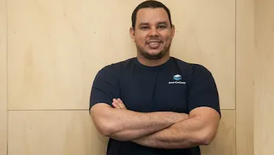 Homem em pé, de braços cruzados, em frente a fundo de madeira.