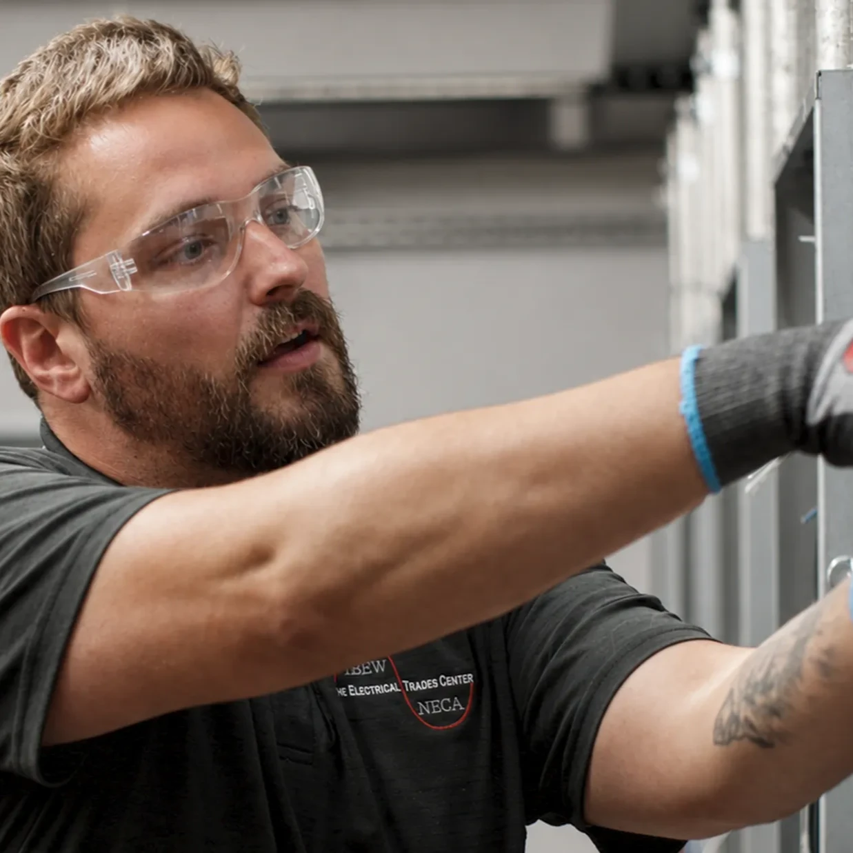 A man with a beard, wearing clear safety glasses, gray work gloves, and a dark short-sleeved polo shirt, works on electrical wiring or panels in an industrial setting.