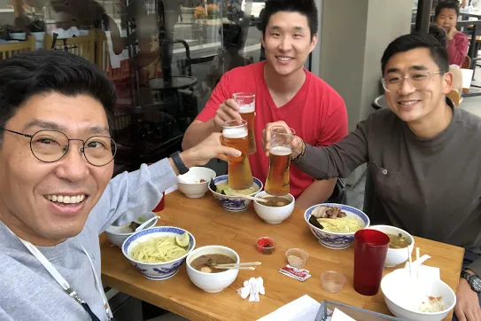 Three men smile looking at the camera while raising their beers together over a table with bowls of noodles.