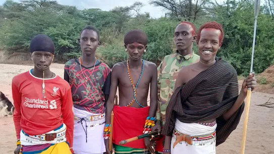 Five young Moran men standing in a dry riverbed in Kenya. They wear a mix of traditional red and patterned shukas, beaded jewelry, and modern clothing, including a red soccer jersey and camouflage jacket. One man holds a traditional spear.