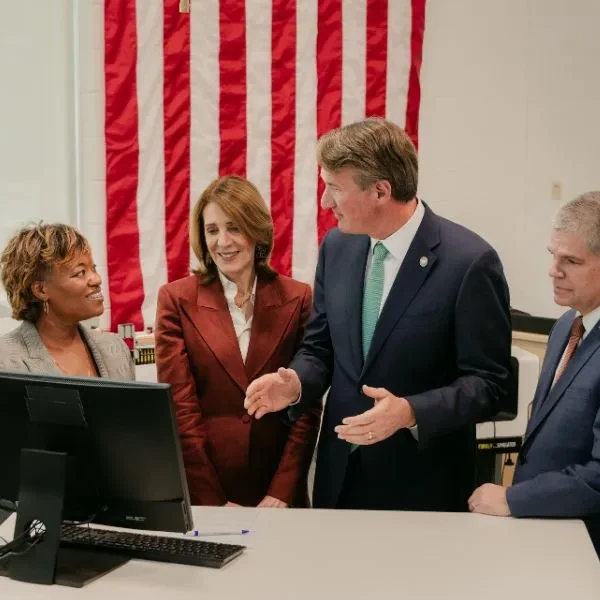 A group of four adults stands together, looking at a demo on a computer screen. They are in a college classroom environment, with lab equipment