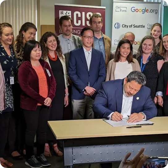 Seattle Mayor Bruce Harrell sits at a desk signing an official document while a group of community partners and representatives from Google.org, CiviForm, and the City of Seattle stand behind him smiling and watching.
