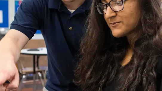 A male teacher in a blue polo shirt leans over to assist a female student wearing glasses as she works on a laptop in a classroom setting.