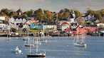 A view across a harbor dotted with sailboats, looking toward a scenic coastal town built on a hill with brightly colored houses, historic buildings, and a church steeple.