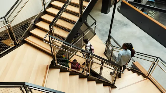 An overhead shot of a modern, multi-level wooden staircase with black industrial railings and wire mesh guards. Several people are seen transitioning between floors: a person in a light blue shirt and dark pants walks down the stairs on the right, while others with backpacks are positioned on the landings and lower flights. The architecture features clean lines, light wood grain, and a polished concrete floor visible at the base of the atrium.