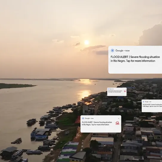 An aerial view of a coastal or riverfront town at sunset, with Google notification cards overlaid on the scene. The main notification card reads: "FLOOD ALERT | Severe flooding situation in Rio Negro. Tap for more information." Additional identical, smaller notification cards are scattered over the cityscape. The background shows a wide river, a sunset reflecting on the water, and densely packed buildings along the shoreline.