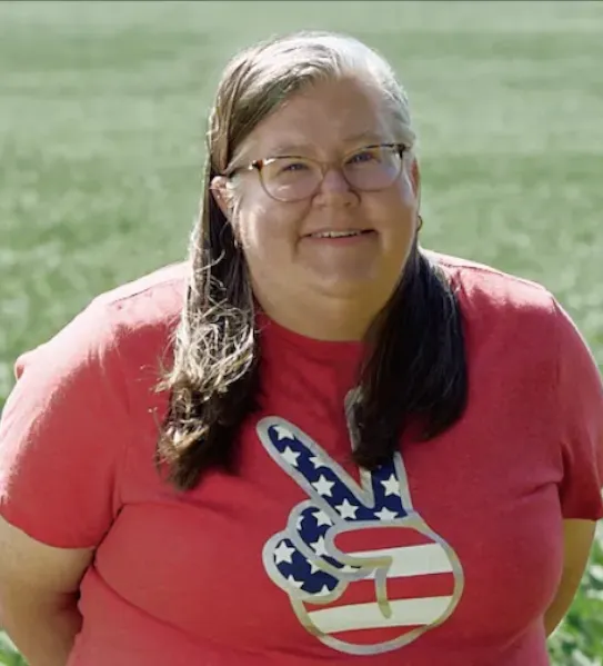 A smiling woman with blonde hair and glasses wearing a red t-shirt with an American flag peace sign logo, standing in a lush green Minnesota field for a Google Gemini educator story.