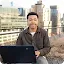 A young man smiling while working on a Google Chromebook on a rooftop with city buildings in the background.