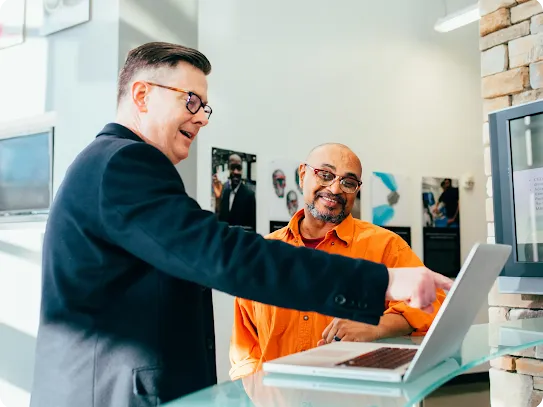 A man in a black blazer and glasses points to a laptop screen while explaining something to a smiling colleague in an orange shirt.
