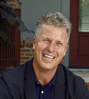 A smiling man with curly blonde hair wearing a dark blazer over a navy blue polo shirt, sitting outdoors.
