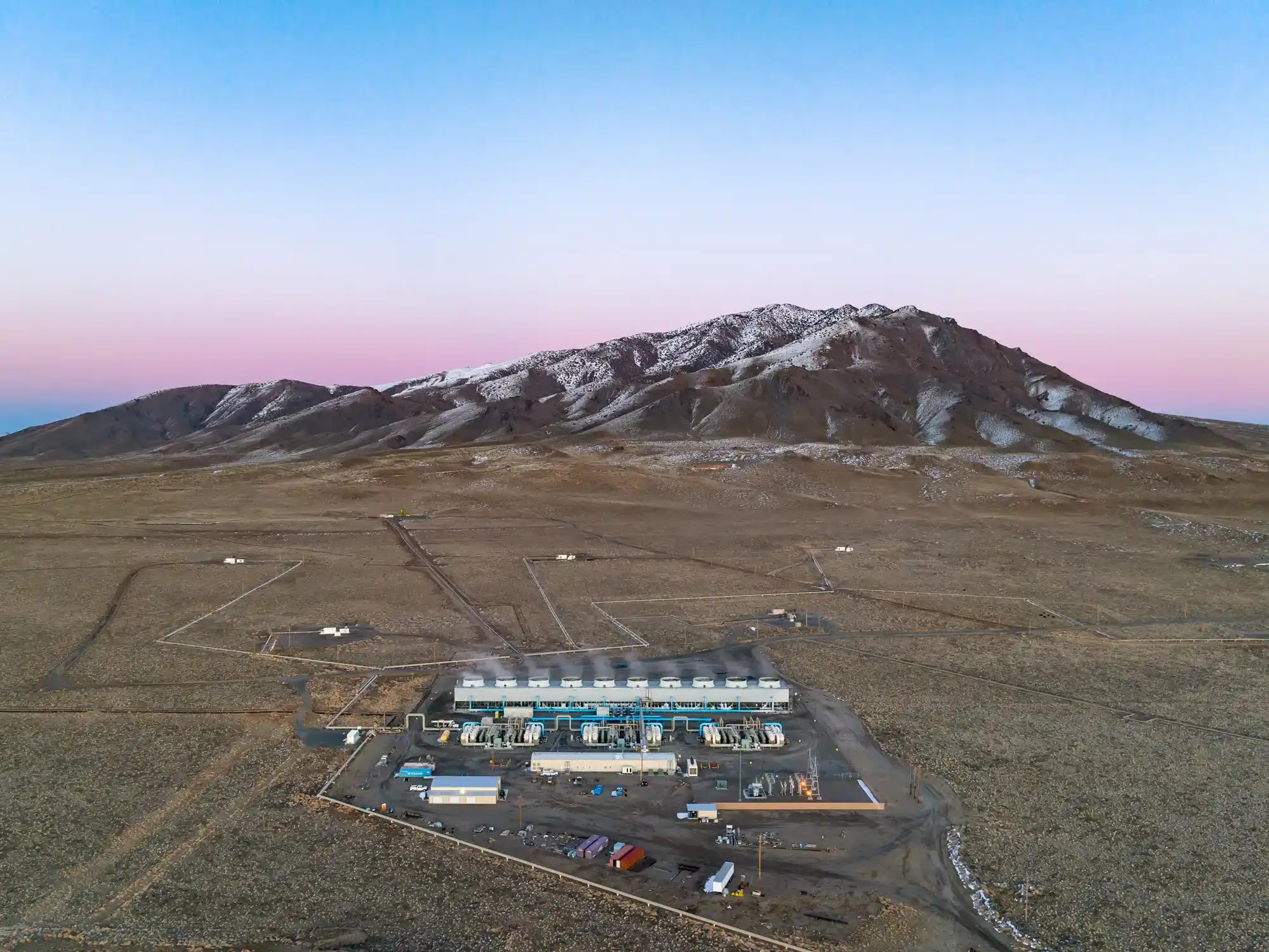 An aerial shot shows a large industrial facility in a barren, arid landscape, with a snow-capped mountain range under a pink and blue sky in the background.
