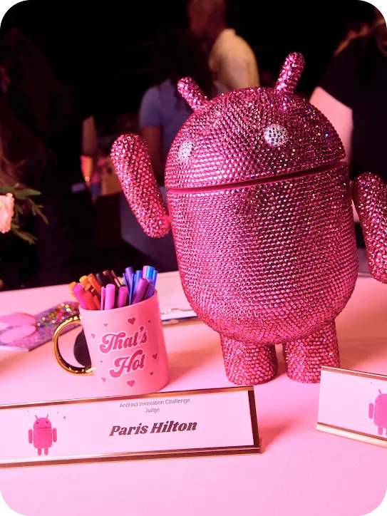 A close-up of a large, bedazzled pink Android mascot on a table next to a pink "That's Hot" mug filled with pens, sitting behind a nameplate identifying Paris Hilton as an Android Innovation Challenge Judge.