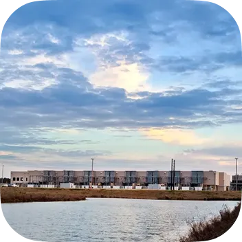 A long, modern industrial building or data center situated behind a calm body of water under a dramatic, cloudy sunset sky. The facility features multiple utility cooling units along its exterior.