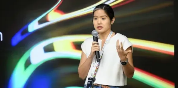 A woman speaking into a microphone on stage against a colorful, abstract background.