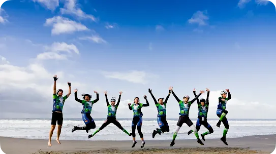 A group of eight cyclists in matching green, blue, and black athletic jerseys and helmets jumping excitedly in the air on a sandy beach. They are lined up side-by-side with arms raised in celebration, set against a background of ocean waves and a bright blue sky with scattered white clouds.