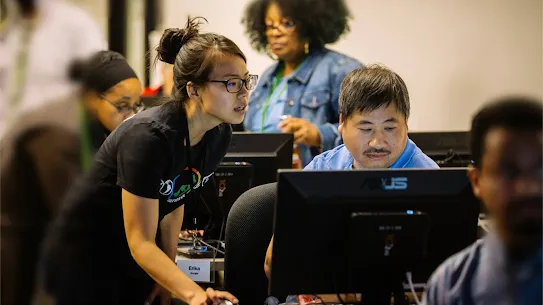 A woman with glasses and her hair in a bun leans over to assist a man sitting at a computer in a crowded workspace. She wears a black t-shirt with a "GoogleServe" logo and an "Erika" name tag. Other people are visible working at computers in the background.