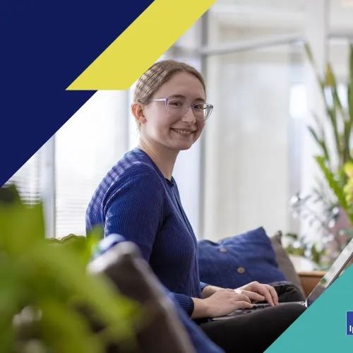 An office worker smiles while sitting on a couch working on a laptop